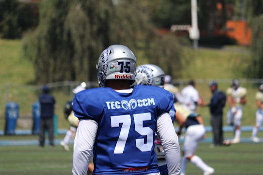 A college football player in uniform on the field, preparing for the game with teammates in the background.