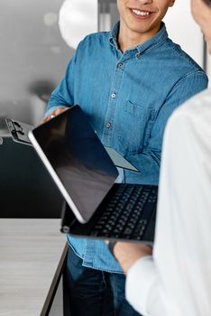 Two adults working together, exchanging a tablet and laptop in an office.